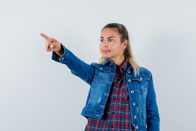 Young lady pointing at something away in shirt, jacket and looking focused , front view.