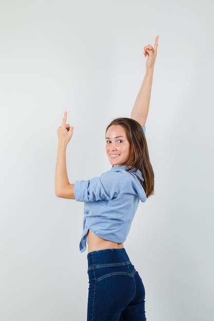 Young lady pointing up by stretching arms in blue shirt, pants and looking cheerful