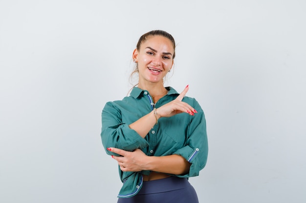 Young lady pointing up in shirt, pants and looking cheerful , front view.