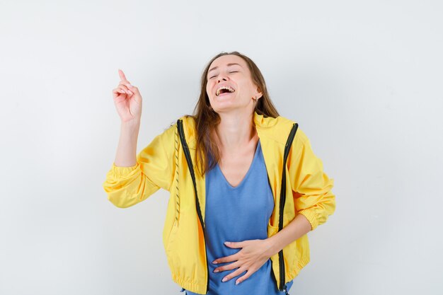 Young lady pointing up in t-shirt, jacket and looking blissful , front view.