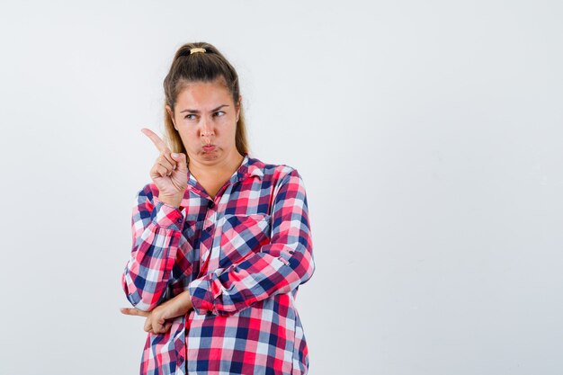 Young lady pointing at upper left corner in checked shirt and looking hesitant , front view.