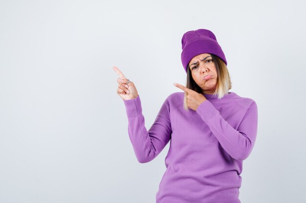 Young lady pointing at upper left corner in purple sweater, beanie and looking disappointed , front view.