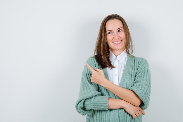 Young lady pointing at upper left corner in shirt, cardigan and looking pleased. front view.