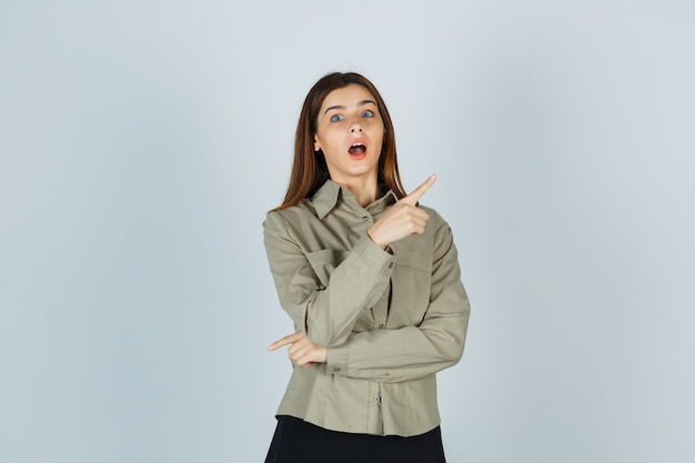 Young lady pointing at upper right corner in shirt, skirt and looking wondered , front view.