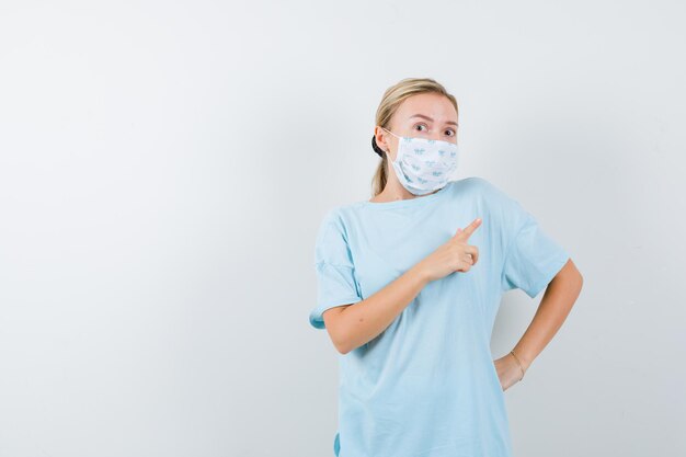 Young lady pointing at upper right corner in t-shirt, mask and looking anxious