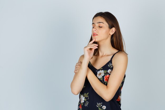 Young lady posing while touching face skin on her chin in blouse and looking delightful , front view.