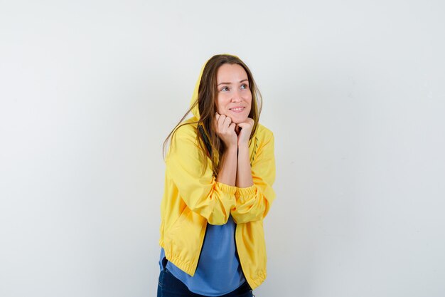 Young lady propping chin on fists in t-shirt, jacket and looking dreamy. front view.