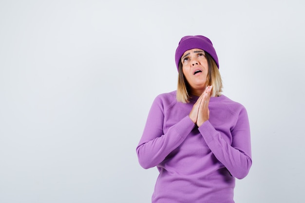 Young lady in purple sweater, beanie with hands in praying gesture and looking hopeful , front view.