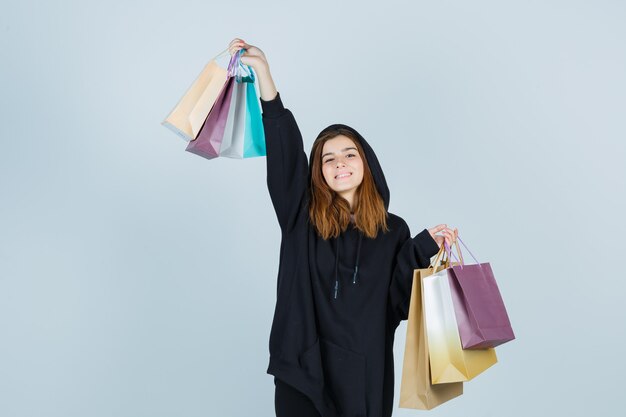 Young lady raising packets while posing in oversized hoodie, pants and looking energetic , front view.