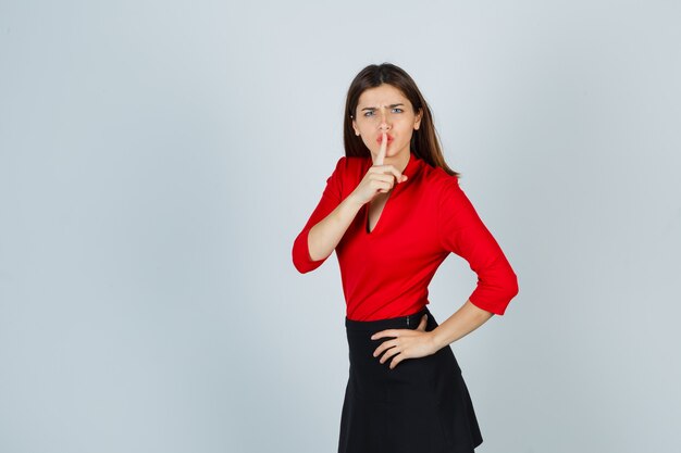 Young lady in red blouse, black skirt showing silence gesture