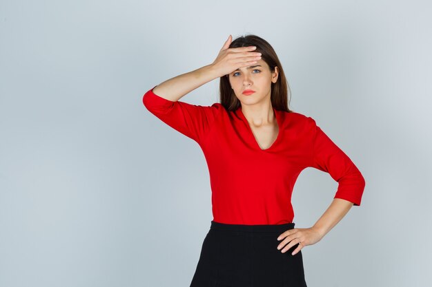 Young lady in red blouse, skirt holding hand on forehead