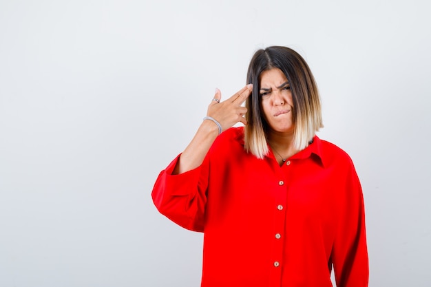 Young lady in red oversize shirt showing gun gesture and looking serious , front view.