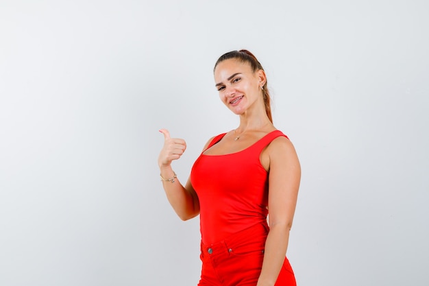 Young lady in red singlet, red trousers showing thumb up and looking pleased , front view.