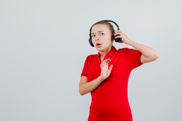 Young lady in red t-shirt taking off headphones with stop gesture and looking scared