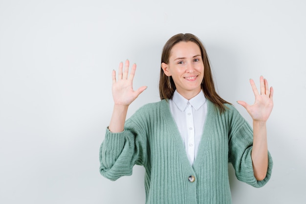 Young lady in shirt, cardigan showing palms and looking joyful , front view.