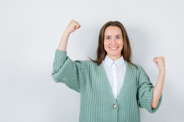 Young lady in shirt, cardigan showing winner gesture and looking lucky , front view.