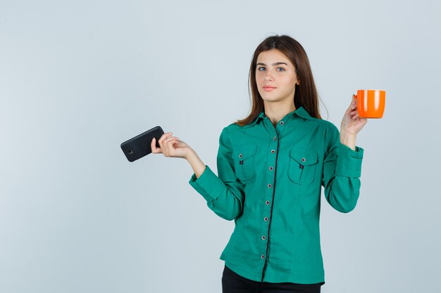 Young lady in shirt holding orange cup of tea and mobile phone and looking confident , front view.