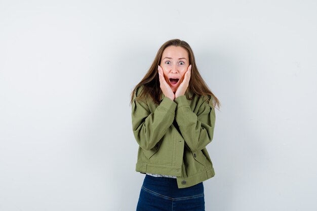 Young lady in shirt, jacket holding hands on cheeks and looking amazed , front view.