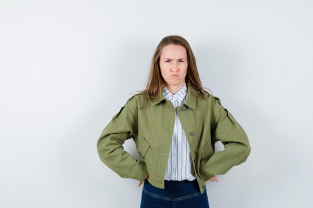 Young lady in shirt, jacket holding hands on waist and looking doubtful, front view.