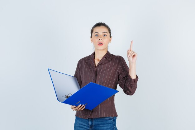 Young lady in shirt, jeans holding folder, pointing up and looking wistful , front view.