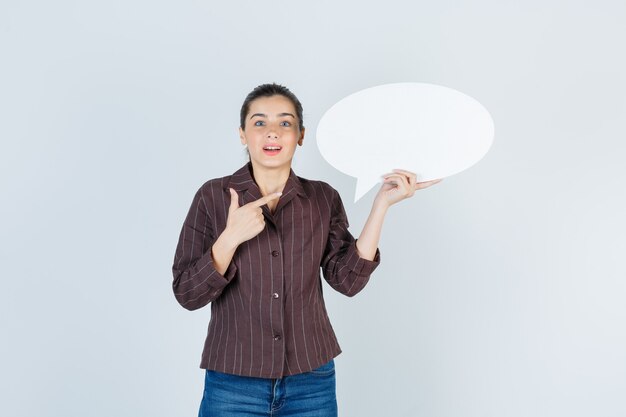 Young lady in shirt, jeans pointing up, keeping paper poster and looking shocked , front view.