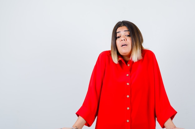 Young lady showing doubt gesture in red oversize shirt and looking perplexed. front view.