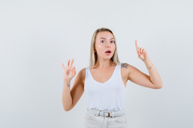 Young lady showing ok gesture while pointing up in white blouse and looking pleased