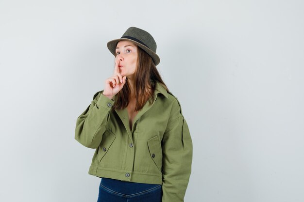 Young lady showing silence gesture in jacket pants hat and looking careful