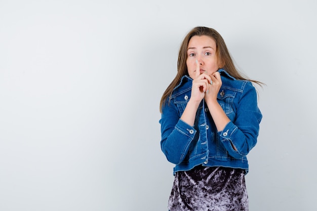 Young lady showing silence gesture, pulling collar on face in dress, denim jacket and looking scared. front view.