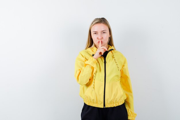 Young lady showing silence gesture in yellow jacket, pants and looking careful. front view.