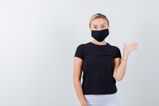 Young lady showing something in t-shirt, pants, medical mask and looking confident