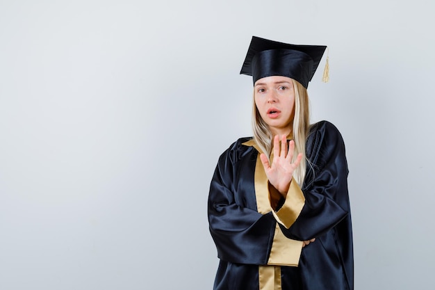 Young lady showing stop gesture in academic dress and looking scared