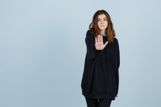 Young lady showing stop gesture in oversized hoodie, pants and looking serious. front view.