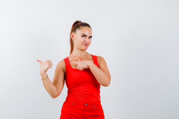 Young lady showing thumb up gesture, pointing to the left side in red singlet, red trousers and looking confident. front view.