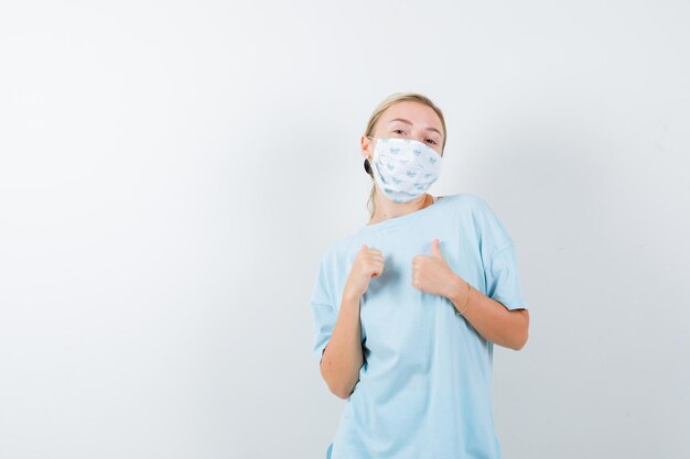 Young lady showing thumbs up in t-shirt, mask and looking pleased