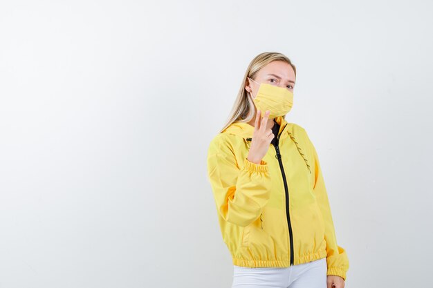 Young lady showing V-sign in jacket, pants, mask and looking confident. front view.