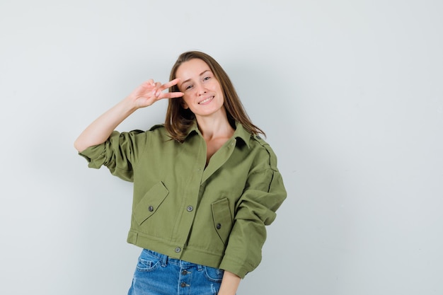 Young lady showing v-sign near eye in green jacket shorts and looking confident  