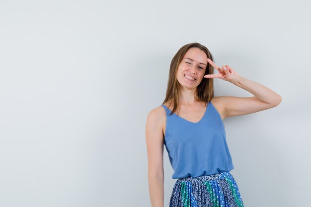 Young lady showing V-sign in singlet, skirt and looking happy , front view.