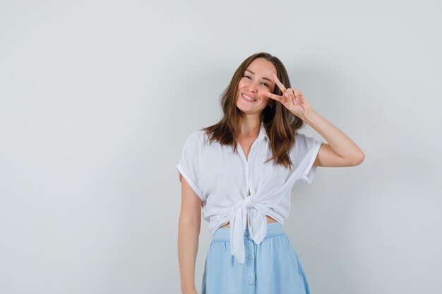 Young lady showing victory gesture in blouse and skirt and looking cheery