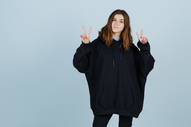 Young lady showing victory sign in oversized hoodie, pants and looking lucky , front view.