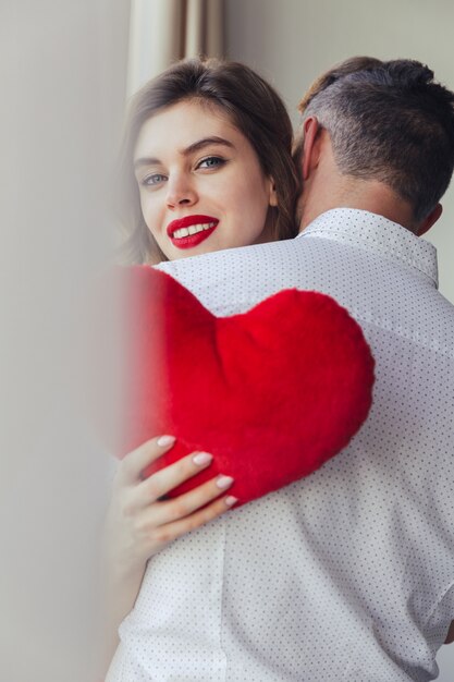 Free Photo young lady smiling and holding heart toy while hug her husband at home