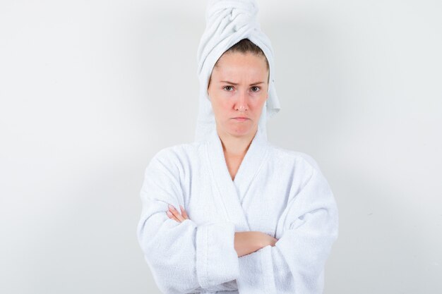 Young lady standing with crossed arms in white bathrobe, towel and looking resentful. front view.