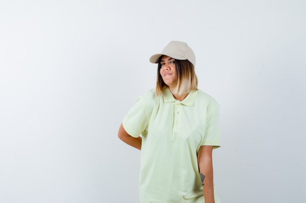 Free photo young lady in t-shirt, cap holding hand behind back while posing and looking pretty , front view.