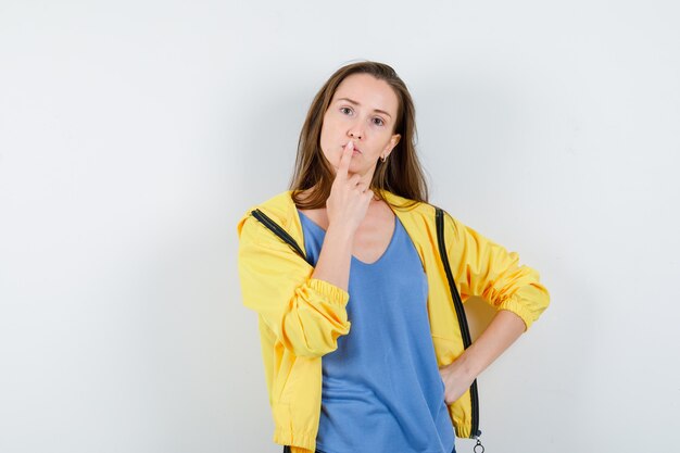 Young lady in t-shirt, jacket holding finger on lips and looking pensive, front view.