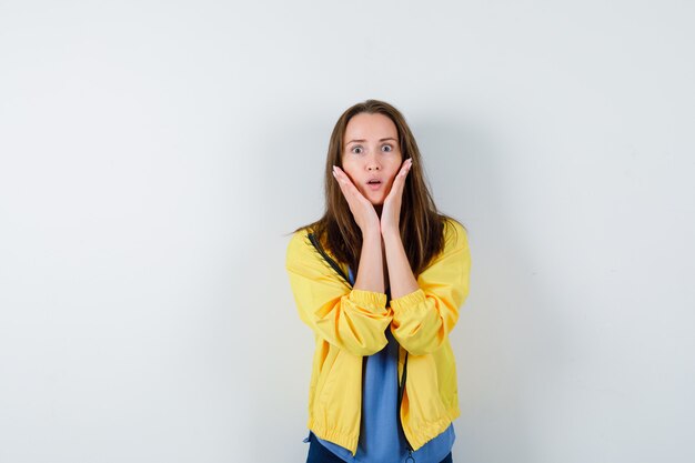 Young lady in t-shirt, jacket holding hands on cheeks and looking surprised, front view.