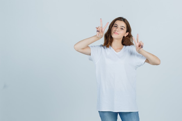 Young lady in t-shirt, jeans showing victory sign and looking cheerful , front view.