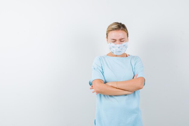 Young lady in t-shirt, mask standing with crossed arms and looking sorrowful