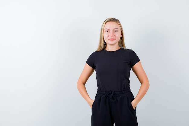 Free photo young lady in t-shirt, pants holding hands in pockets and looking merry , front view.