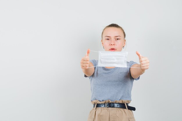 Young lady in t-shirt and pants showing medical mask and looking careful