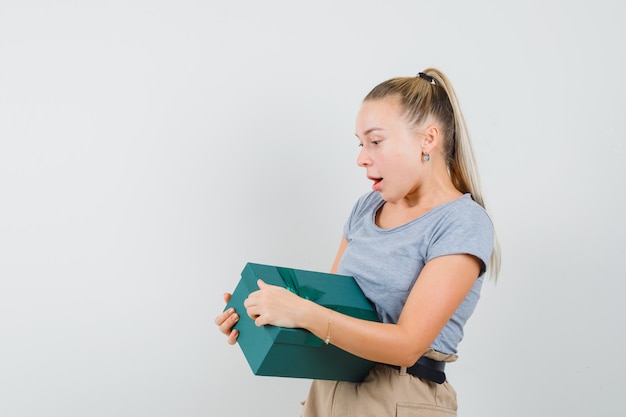 Young lady in t-shirt and pants trying to open present box and looking curious,